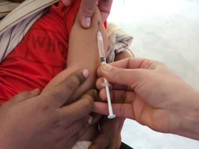 Criança recebe uma dose de vacina aplicada por profissional de saúde em uma unidade clínica.