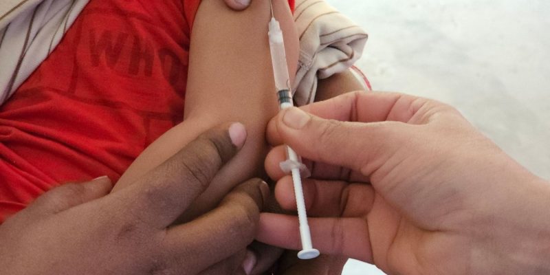 Criança recebe uma dose de vacina aplicada por profissional de saúde em uma unidade clínica.