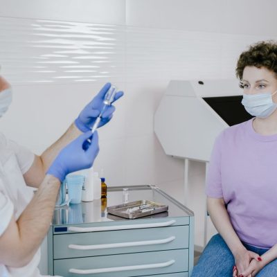 Profissional de saúde aplica uma vacina em um paciente em ambiente clínico; ambos usam máscaras.