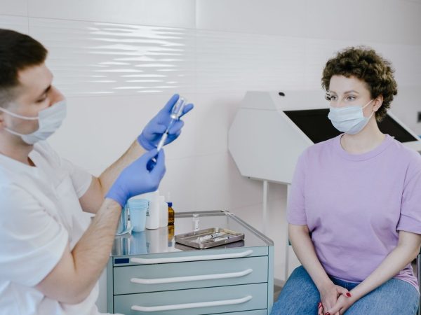Profissional de saúde aplica uma vacina em um paciente em ambiente clínico; ambos usam máscaras.