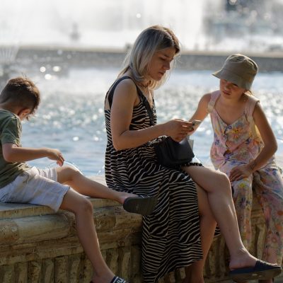 Família com crianças em um parque, enquanto um adulto utiliza um smartphone próximo a uma fonte.