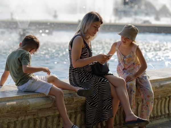 Família com crianças em um parque, enquanto um adulto utiliza um smartphone próximo a uma fonte.