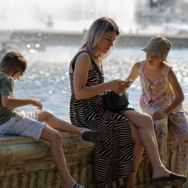 Família com crianças em um parque, enquanto um adulto utiliza um smartphone próximo a uma fonte.