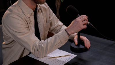Profissional prepara um podcast em estúdio, diante de um microfone e anotações.